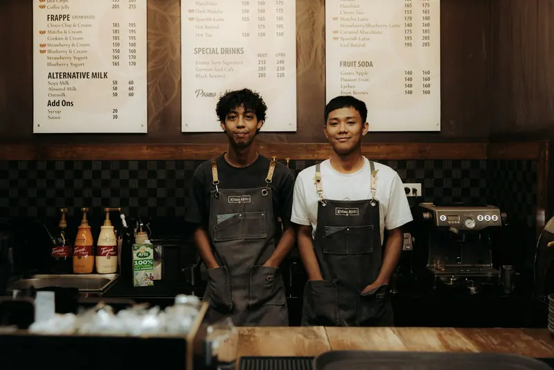 Two baristas in aprons behind the counter, under the menu boards.