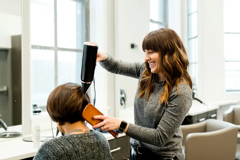 A stylist laughing as she blow-dries a client’s hair.